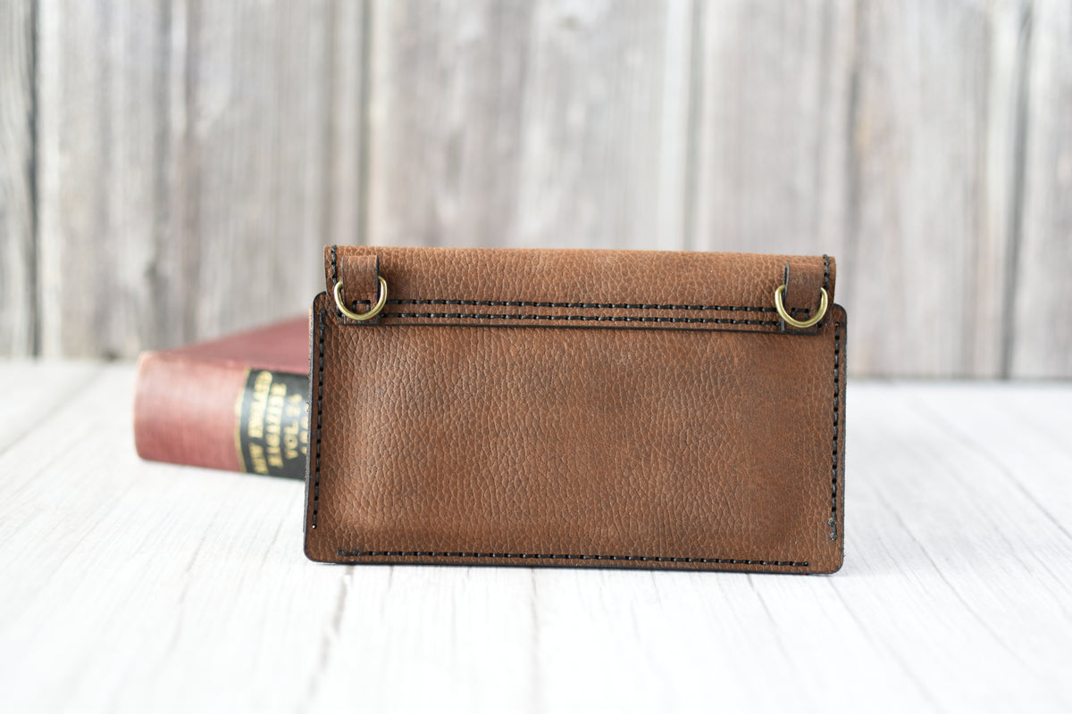 Brown leather cell phone purse on a wooden surface with a book in the background