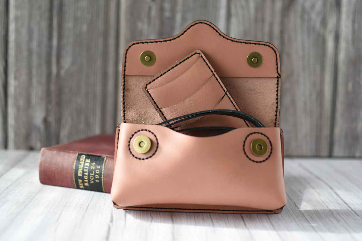 Brown leather cell phone purse on a wooden surface with a book in the background with compact wallet and black strap