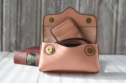 Brown leather cell phone purse on a wooden surface with a book in the background with compact wallet and black strap