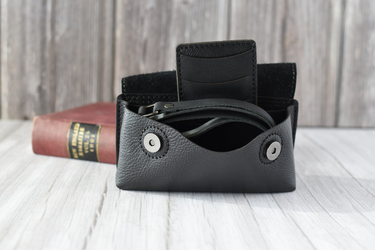 Black leather cell phone purse on a wooden surface with a book in the background with compact wallet and black strap