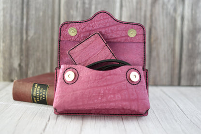 Pink leather cell phone purse on a wooden surface with a book in the background with compact wallet and strap